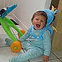Ayden a rejoint le concours — aidez-le/la à gagner de superbes lots ! baby, child, toddler, laughing, blue_onesie, dinosaur_backpack, toy_walker, green_toy, orange_wheels, indoor, tiled_floor, happy, cute, smiling, sitting, playful, home, young_child, floor, fun