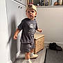 toddler, child, cap, clothing, shorts, socks, shoes, standing, indoor, flooring, rug, wicker_stool, cabinet, photograph, frame, decor, person, expression, curious, hat