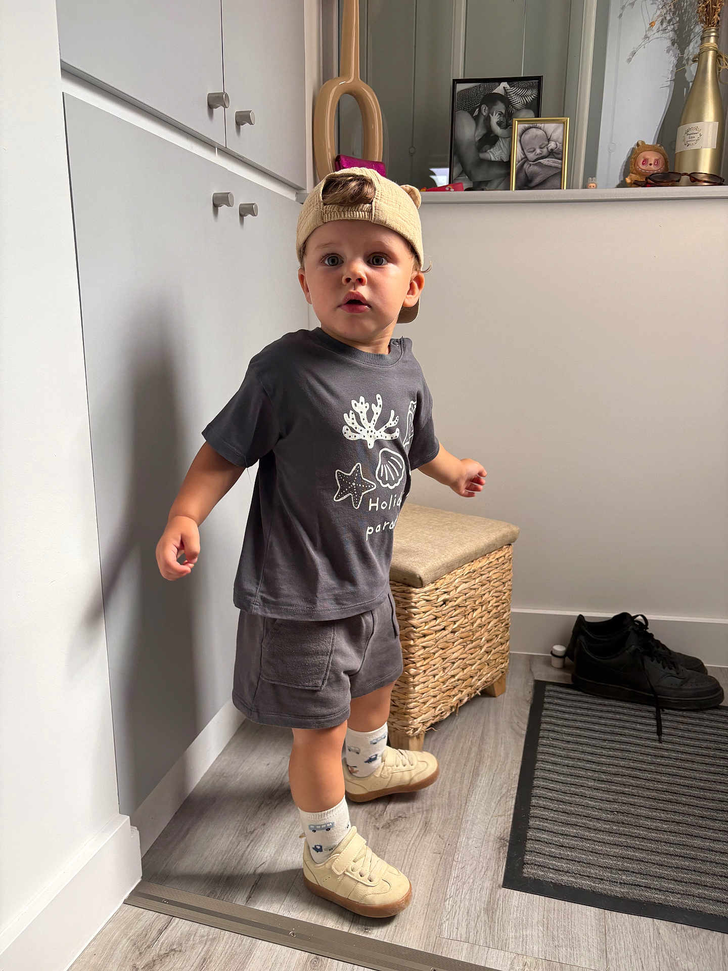 Mylo participe au concours pour gagner de l'argent avec cette photo : toddler, child, cap, clothing, shorts, socks, shoes, standing, indoor, flooring, rug, wicker_stool, cabinet, photograph, frame, decor, person, expression, curious, hat