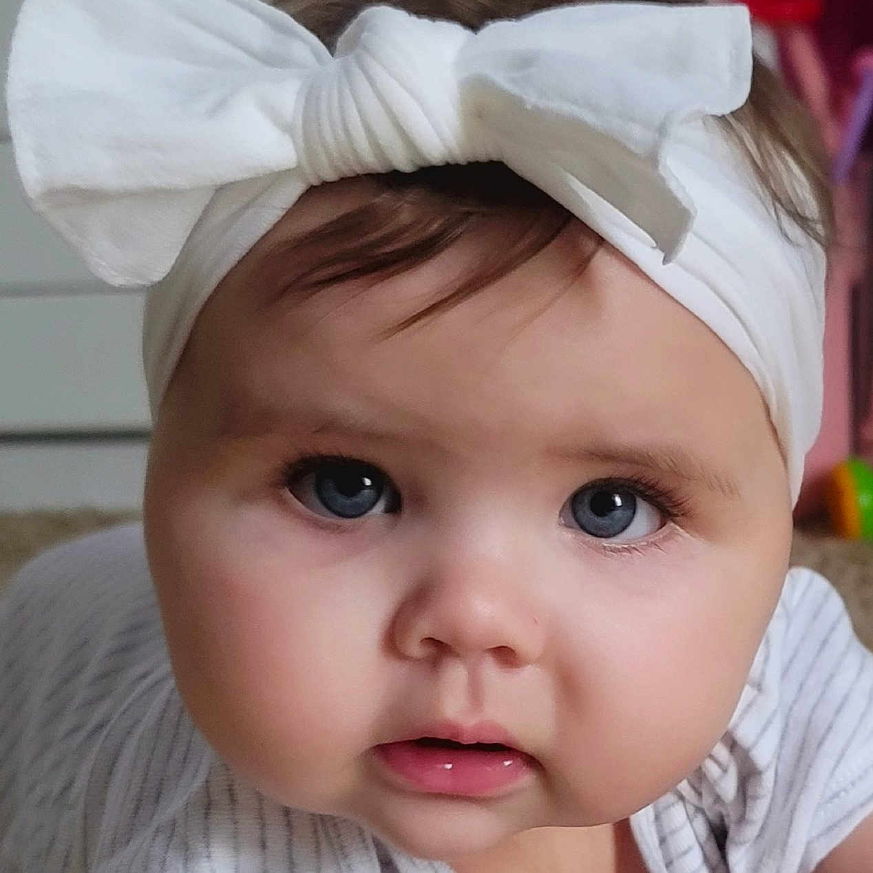 Whitley is registered to the contest to win money with this photo: baby, child, infant, portrait, face, eyes, wide_eyed, white_bow, headband, striped_clothing, white_clothing, sitting, indoor, home, carpet, furniture, close_up, looking_at_camera, cute, photography