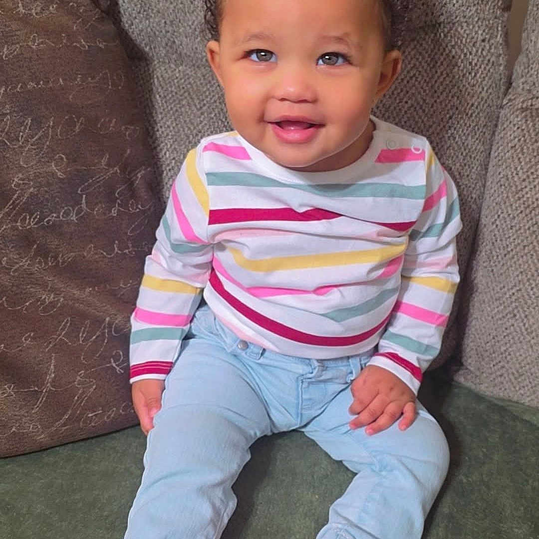 Kaliah joined the competition — help win amazing prizes! child, couch, cozy, cute, face, fashion, feet, hair, happy, indoors, light_blue_jeans, pink_bows, pink_slippers, playful, portrait, sitting, smiling, striped_shirt, toddler, young_child