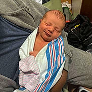 Atticus is registered to the contest to win money with this photo: newborn, baby, infant, wrapped, blanket, striped_blanket, sleeping, person, clothing, sweatpants, hoodie, hand, indoor, floor, bag, casual, resting, cozy, smile, human