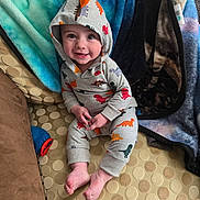Jordan is registered to the contest to win money with this photo: baby, blanket, child, colorful_clothing, couch, cozy, cute, dinosaur_hoodie, feet, happy, hoodie, indoors, infant, patterned_fabric, person, playful, portrait, seated, smiling, soft_textiles