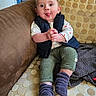 baby, child, couch, clothing, vest, pants, socks, tongue, hands, face, indoor, pattern, furniture, playful, smile, person, sitting, expression, cute, home