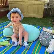 Jordy joined the competition — help win amazing prizes! baby, child, outdoor, grass, blanket, hat, pillow, book, smiling, sitting, lying_down, fence, toy, clothing, infant, cute, playful, daylight, relaxing, portrait