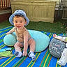 baby, child, outdoor, grass, blanket, hat, pillow, book, smiling, sitting, lying_down, fence, toy, clothing, infant, cute, playful, daylight, relaxing, portrait