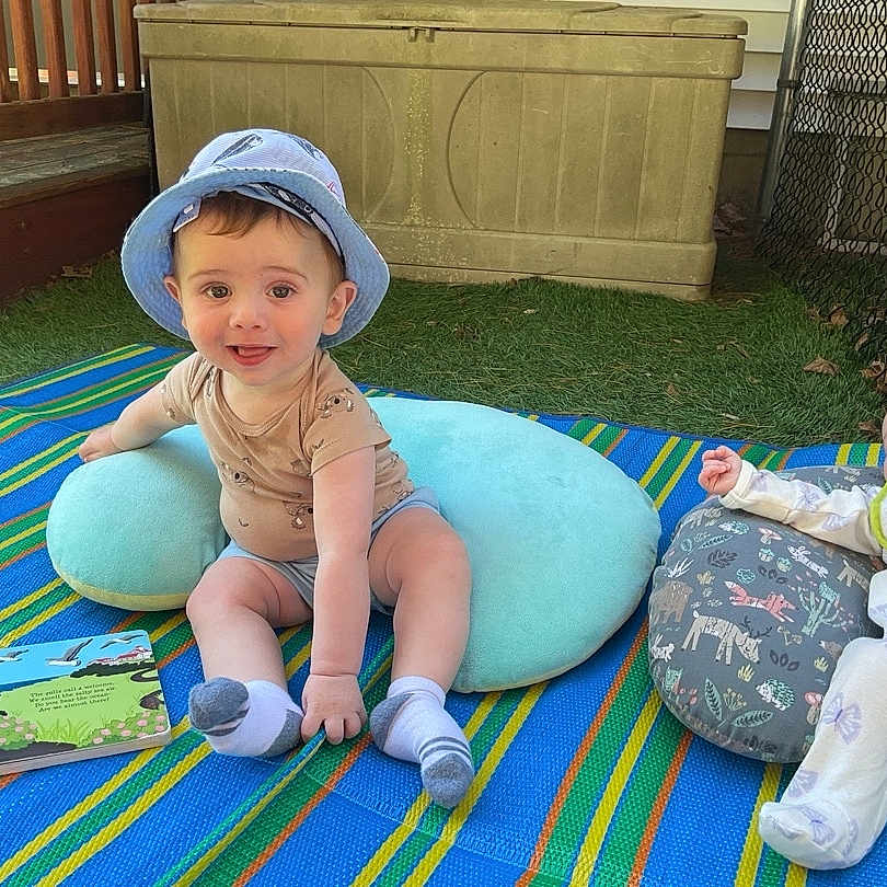 Jordy joined the competition — help win amazing prizes! baby, child, outdoor, grass, blanket, hat, pillow, book, smiling, sitting, lying_down, fence, toy, clothing, infant, cute, playful, daylight, relaxing, portrait