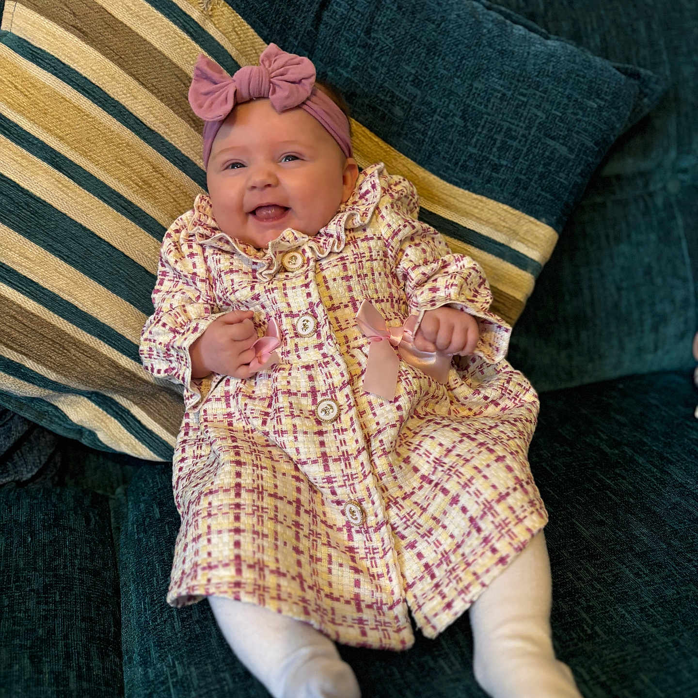 Elda-Rose is registered to the contest to win money with this photo: baby, bow, child, coat, couch, cozy, cute, happy, headband, indoors, infant, patterned_clothing, pillows, pink, portrait, seated, smiling, soft_furniture, striped_pillow, white_tights