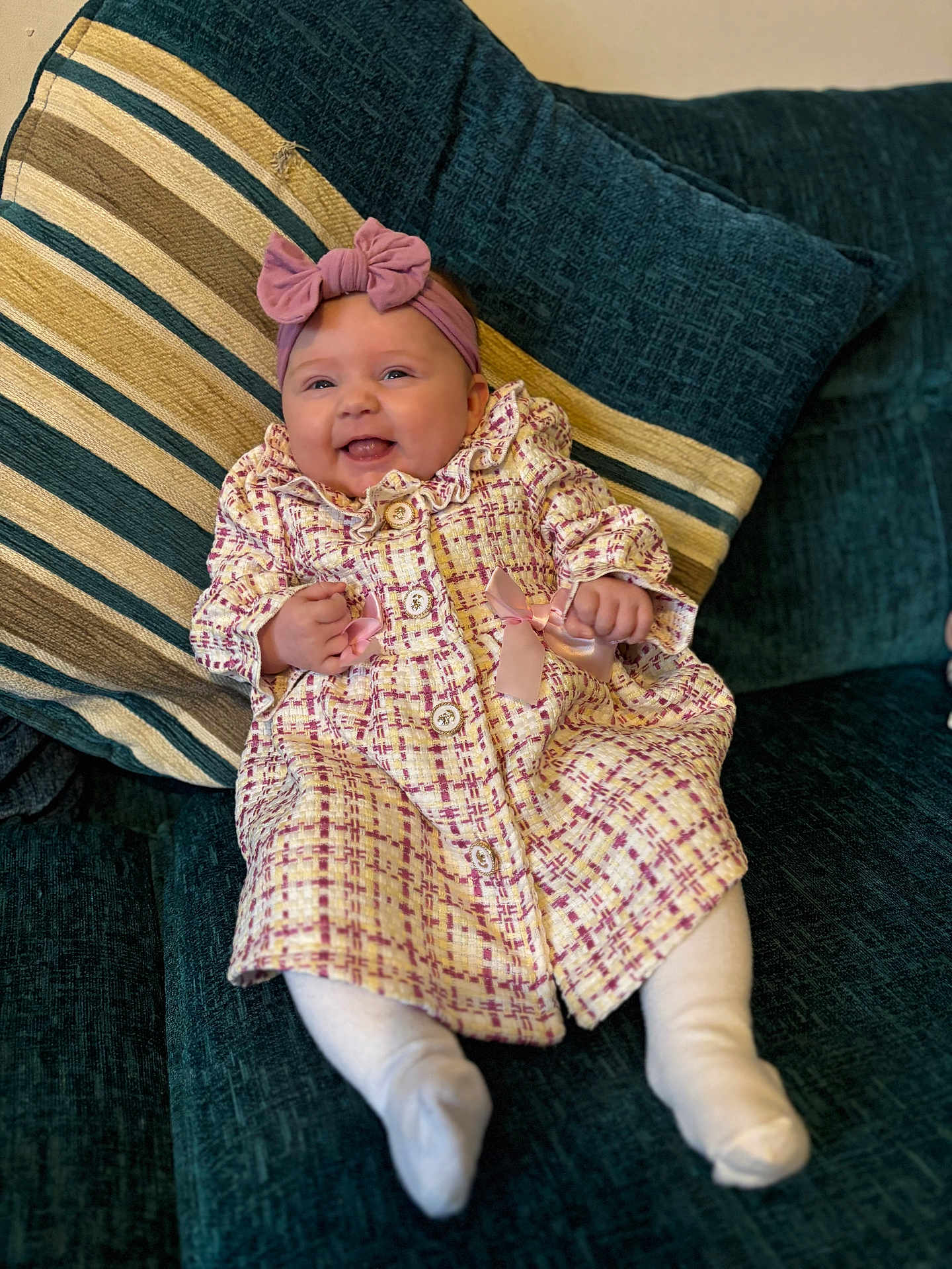 Elda-Rose is registered to the contest to win money with this photo: baby, bow, child, coat, couch, cozy, cute, happy, headband, indoors, infant, patterned_clothing, pillows, pink, portrait, seated, smiling, soft_furniture, striped_pillow, white_tights