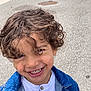 child, smiling, curly_hair, denim_jacket, white_shirt, outdoor, pavement, manhole_cover, face, happy, person, casual_clothing, closeup, portrait, young_child, cute, daylight, boy, emotion, fun