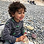 child, curly_hair, smiling, ice_cream, cup, spoon, pebble_beach, rocks, casual_clothing, gray_sweater, blue_pants, outdoor, daytime, happy, sitting, food, dessert, stone_wall, people_in_background, relaxed