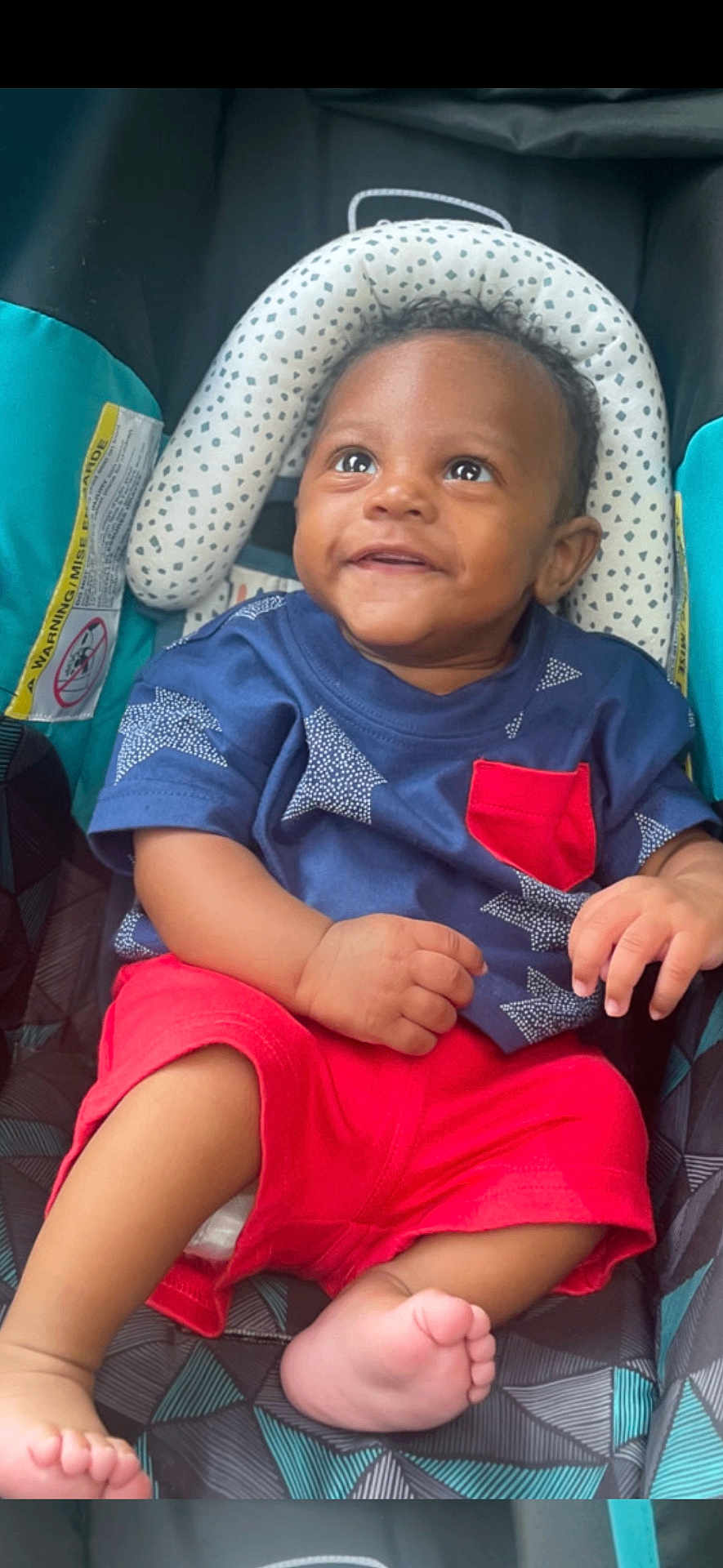 Denim is registered to the contest to win money with this photo: baby, child, smiling, blue_shirt, red_shorts, baby_seat, patterned, curly_hair, happy, infant, cute, portrait, sitting, indoors, small_hands, barefoot, bright_colors, closeup, young, person