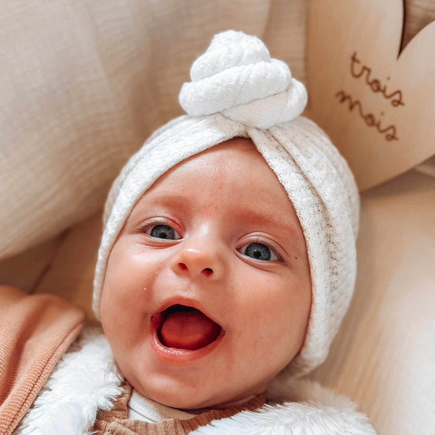 Emma participe au concours pour gagner de l'argent avec cette photo : baby, blanket, blue_eyes, cozy, cute, face, fur_vest, happy, headwear, heart_shape, indoors, infant, neutral_colors, open_mouth, portrait, smiling, soft_texture, white_hat, wooden_sign, young_child