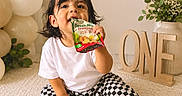Jasper is registered to the contest to win money with this photo: child, toddler, birthday, cake, balloons, fruit_snack, snack_pouch, sitting, smiling, portrait, indoor, celebration, one_year_old, white_shirt, checkered_pants, socks, carpet, decorations, greenery, wooden_sign