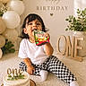child, toddler, birthday, cake, balloons, fruit_snack, snack_pouch, sitting, smiling, portrait, indoor, celebration, one_year_old, white_shirt, checkered_pants, socks, carpet, decorations, greenery, wooden_sign
