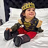 child, toddler, baby, crown, costume, royal, sitting, bed, bedroom, portrait, smiling, happy, face, person, black_clothing, red_pants, boots, pillow, headboard, indoor
