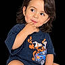 child, toddler, portrait, sitting, smiling, curious, face, eyes, dark_hair, brown_hair, short_sleeves, t_shirt, cartoon_graphic, colorful_clothing, hand, finger, black_background, studio_photo, close_up, safe