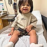 child, toddler, smiling, happy, portrait, sitting, medical_room, examination_bed, healthcare, doctor_office, medical_equipment, stethoscope, blood_pressure_cuff, hospital, indoor, shirt, shorts, socks, long_hair, playful