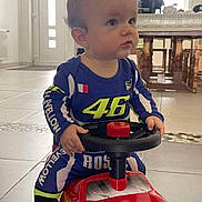 Lévy a rejoint le concours — aidez-le/la à gagner de superbes lots ! baby, child, toy_car, racing_suit, indoor, floor_tiles, steering_wheel, red, blue, yellow, person, small_vehicle, expression, light, window, door, table, chair, footwear, play