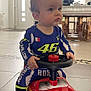 baby, child, toy_car, racing_suit, indoor, floor_tiles, steering_wheel, red, blue, yellow, person, small_vehicle, expression, light, window, door, table, chair, footwear, play