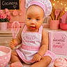 baby, child, infant, chef_hat, apron, pink, kitchen, baking, whisk, cupcake, mixing_bowl, rolling_pin, cookie_cutter, pastry, dessert, sitting, portrait, smiling, indoor, cooking