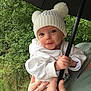 baby, child, white_hat, pom_poms, umbrella, hands, adult_hand, green_jacket, outdoor, nature, foliage, cute, smiling, portrait, person, clothing, headwear, holding, close_up, face