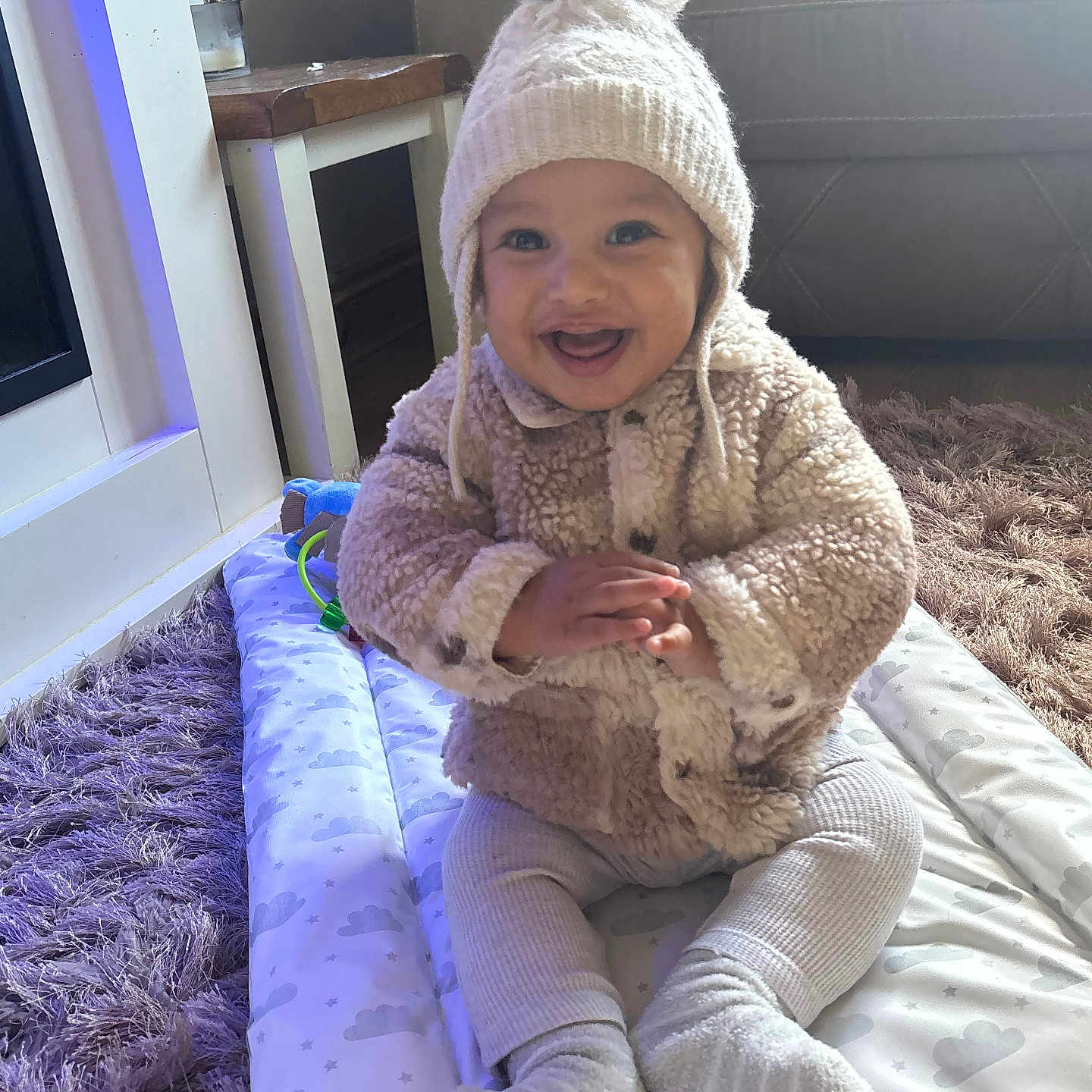 Kayson-Jay is registered to the contest to win money with this photo: baby, child, cloud_pattern, couch, furniture, fuzzy_jacket, happy, hat, home, indoor, knitted_hat, leggings, play_mat, pom_pom, purple_rug, rug, sitting, smiling, socks, table