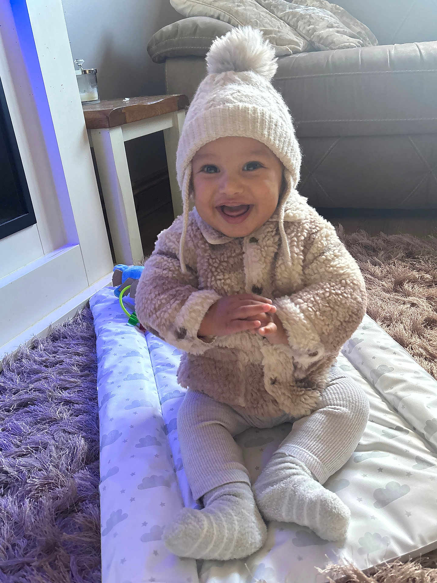 Kayson-Jay is registered to the contest to win money with this photo: baby, child, smiling, hat, pom_pom, fuzzy_jacket, knitted_hat, play_mat, cloud_pattern, socks, leggings, rug, purple_rug, indoor, home, furniture, table, couch, happy, sitting