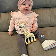 Luna participe au concours pour gagner de l'argent avec cette photo : baby, child, smiling, toy, giraffe, headband, flower, pink_clothing, pants, white_shoes, couch, indoor, cute, happy, sitting, infant, playful, soft_toy, footwear, portrait