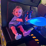 Ayden is registered to the contest to win money with this photo: toddler, child, arcade_game, gaming, controller, seat, shoes, curly_hair, indoor, play, fun, colorful, lighting, entertainment, casual_clothing, blue_shirt, shorts, smiling, young_child, hands