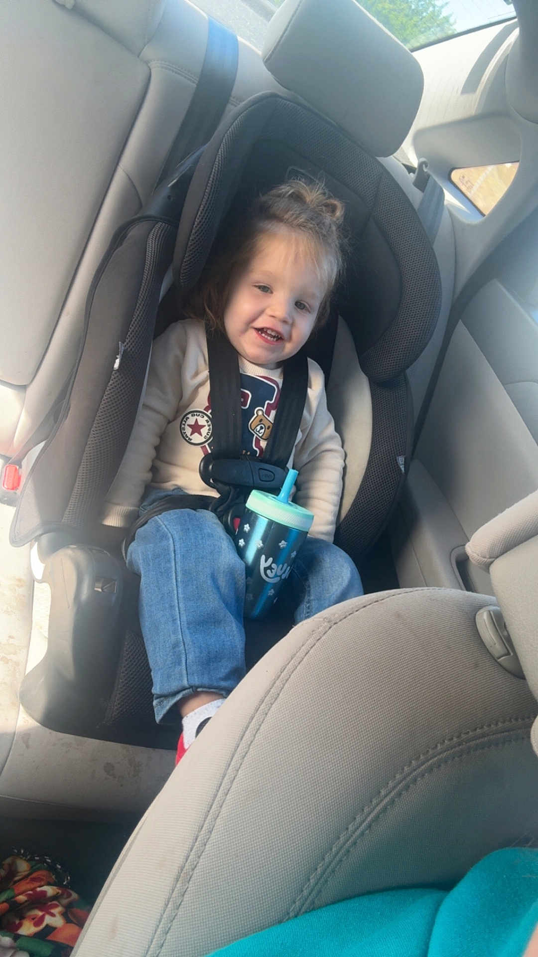 Ayden is registered to the contest to win money with this photo: toddler, child, car_seat, sippy_cup, jeans, sweater, seatbelt, car_interior, smiling, happy, seat, window, sunlight, casual_clothing, person, young_child, hair_tied_up, footwear, safety, daytime