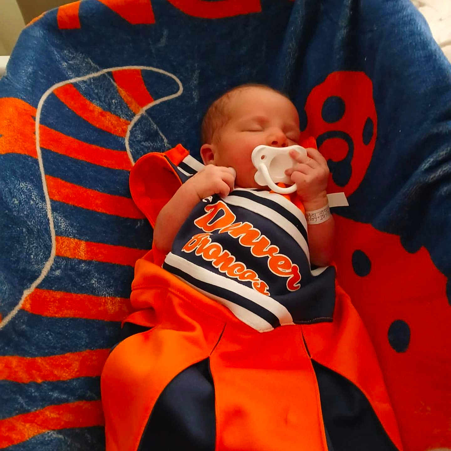 Celine is registered to the contest to win money with this photo: baby, blanket, blue, chair, clothing, cozy, cute, denver_broncos, indoor, infant, newborn, nursery, orange, pacifier, person, portrait, resting, sleeping, soft_lighting, sports_team