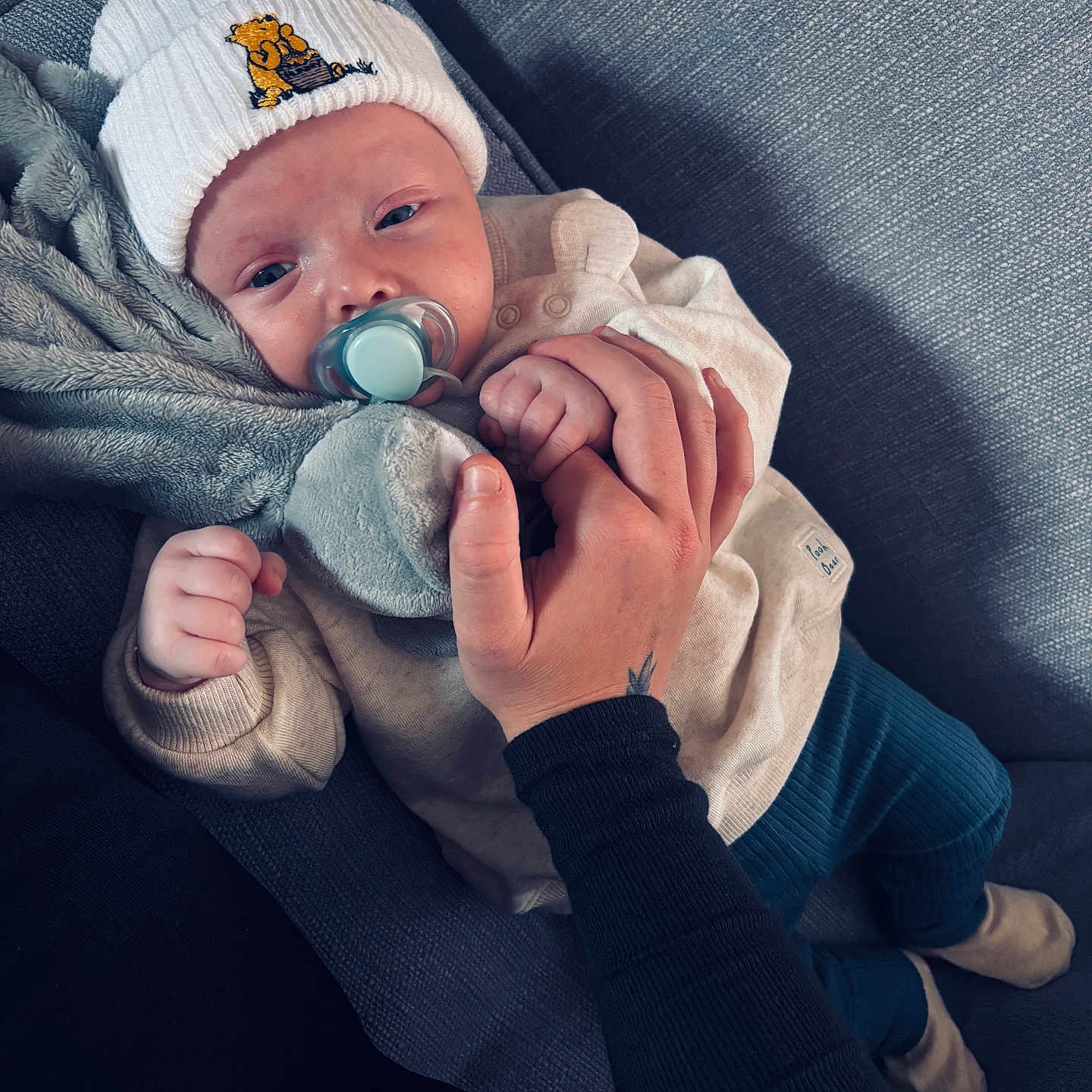 Louie is registered to the contest to win money with this photo: baby, blue_pants, clothing, comfort, couch, cute, fabric, hand, hat, holding, indoor, infant, pacifier, person, relaxed, sitting, skin, soft, toy, warm