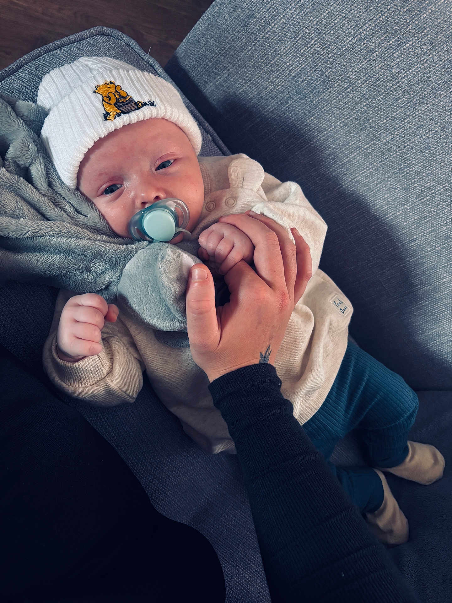 Louie is registered to the contest to win money with this photo: baby, pacifier, hand, hat, clothing, couch, fabric, person, infant, sitting, comfort, indoor, cute, warm, soft, relaxed, holding, skin, blue_pants, toy