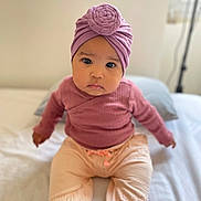 Inaya a rejoint le concours — aidez-le/la à gagner de superbes lots ! baby, infant, toddler, child, headwrap, turban, knit_hat, pink_clothing, mauve_top, beige_pants, pink_socks, big_eyes, sitting, bed, portrait, adorable, indoor, soft_lighting, shallow_depth_of_field, looking_at_camera