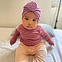 baby, infant, toddler, child, headwrap, turban, knit_hat, pink_clothing, mauve_top, beige_pants, pink_socks, big_eyes, sitting, bed, portrait, adorable, indoor, soft_lighting, shallow_depth_of_field, looking_at_camera