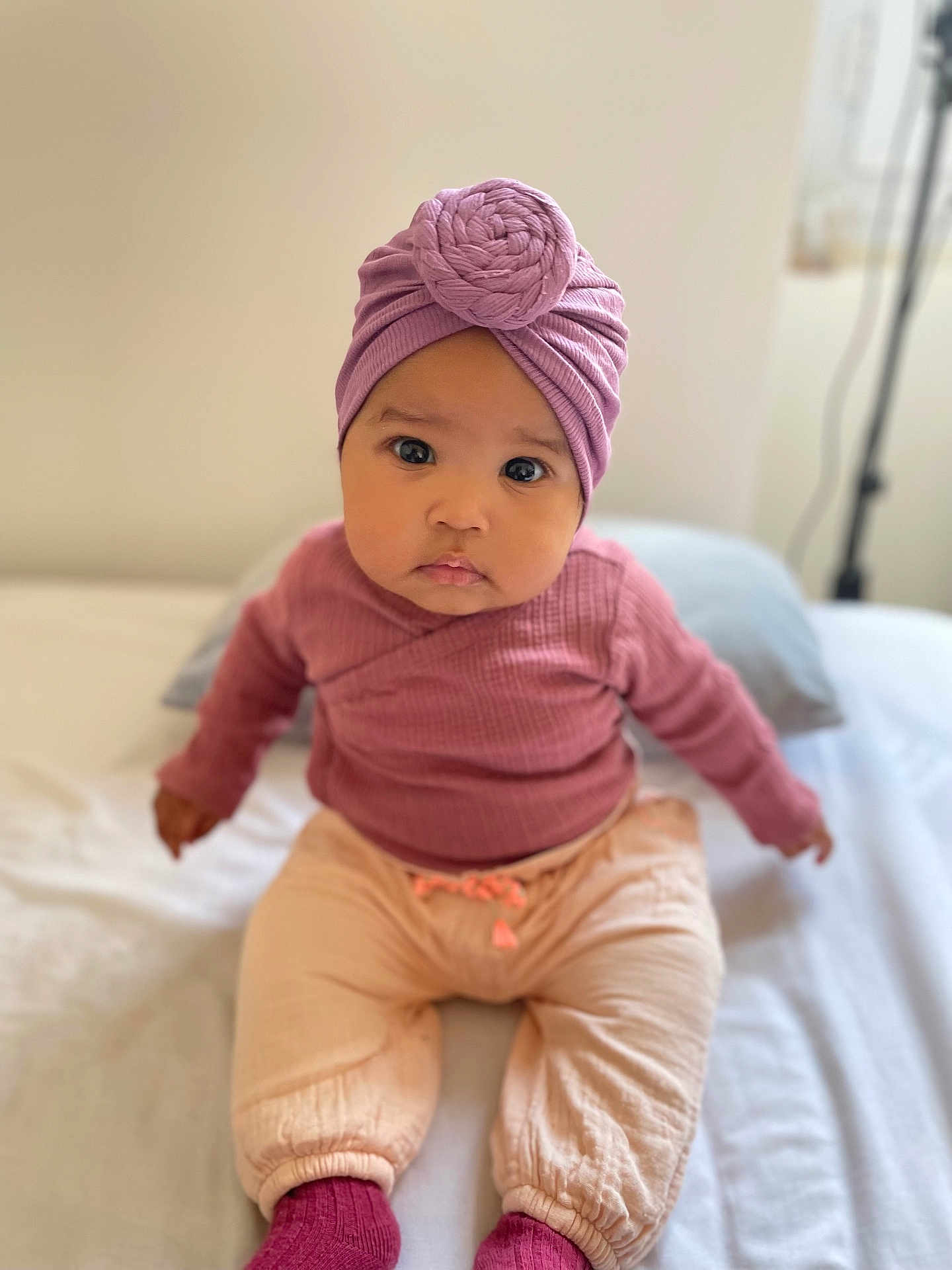 Inaya a rejoint le concours — aidez-le/la à gagner de superbes lots ! baby, infant, toddler, child, headwrap, turban, knit_hat, pink_clothing, mauve_top, beige_pants, pink_socks, big_eyes, sitting, bed, portrait, adorable, indoor, soft_lighting, shallow_depth_of_field, looking_at_camera