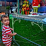 background, child, christmas_decorations, decoration, fence, festive, gift_boxes, grass, green_grass, grinch_figure, holiday_lights, holiday_scene, outdoor, pacifier, person, red_and_white_striped_clothing, reindeer_lights, summer_clothing, toddler, toy