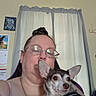 Groot is registered to the contest to win money with this photo: adult, animal, background, cuddling, curtain, dog, eyeglasses, face, female, indoors, jewelry, person, pet, portrait, room, shirt, sitting, small_dog, window, woman