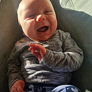George is registered to the contest to win money with this photo: baby, smiling, happy, child, infant, clothing, striped_shirt, patterned_pants, sitting, cushion, chair, indoor, sunlight, shadow, cute, portrait, person, hand, face, cozy