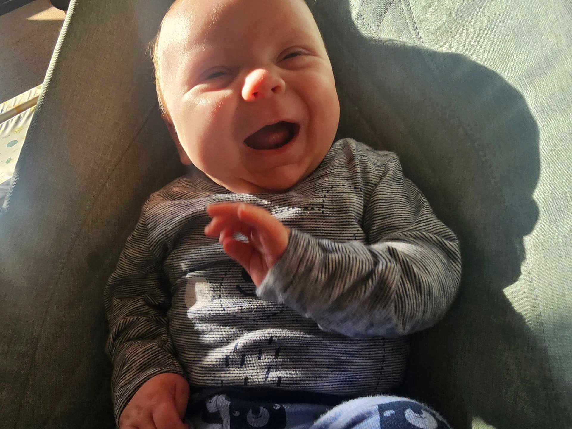 George is registered to the contest to win money with this photo: baby, smiling, happy, child, infant, clothing, striped_shirt, patterned_pants, sitting, cushion, chair, indoor, sunlight, shadow, cute, portrait, person, hand, face, cozy