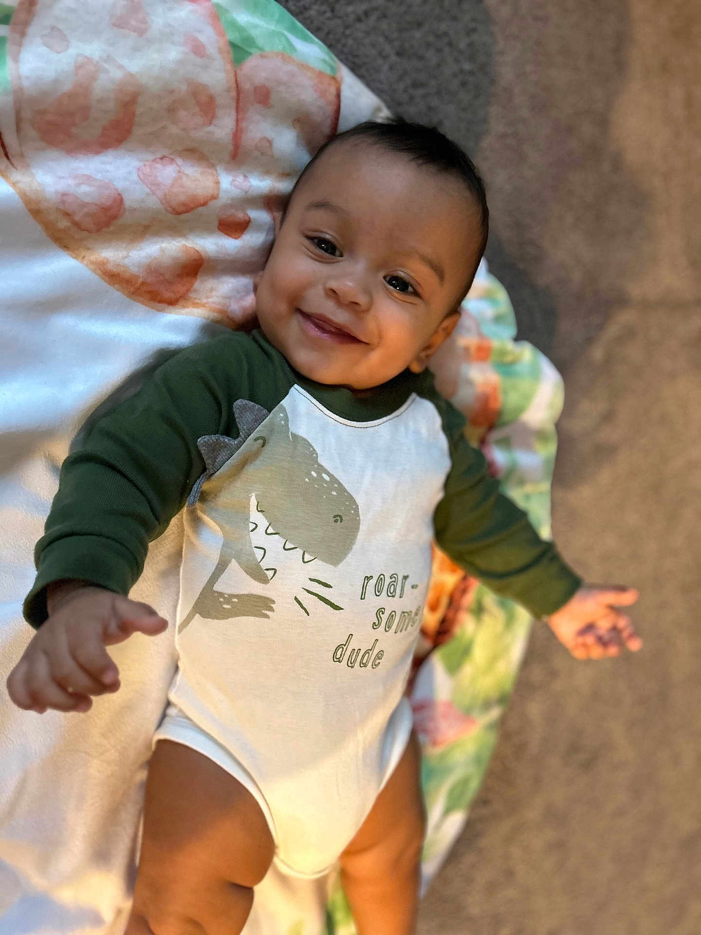 Dennis is registered to the contest to win money with this photo: baby, infant, smile, onesie, dinosaur_print, blanket, carpet, child, cute, happy, lying_down, indoor, reaching, toddler, clothing, portrait, skin, head, face, young