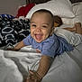 Dennis is registered to the contest to win money with this photo: baby, smiling, bed, pillow, blanket, happy, infant, child, indoor, person, cute, clothing, blue_shirt, white_striped_pants, face, hand, lying_down, expression, joyful, cozy
