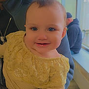 Alaska is registered to the contest to win money with this photo: adult, baby, background_person, beanie, cheeks, closeup, eyes, hand, happy, indoor, infant, lace_dress, natural_light, neck_pillow, parent, portrait, seat, smiling, sweater, window