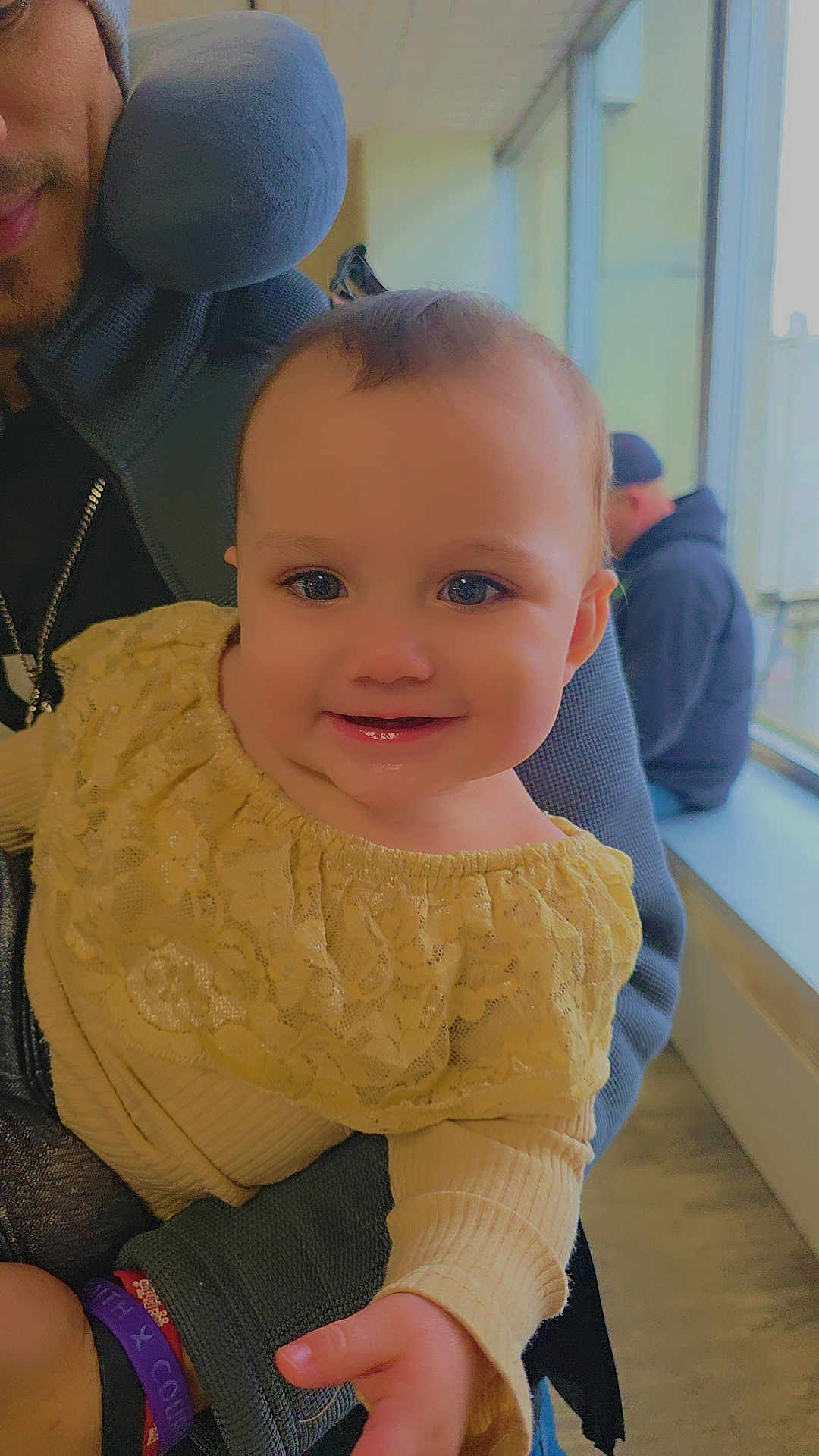 Alaska is registered to the contest to win money with this photo: adult, baby, background_person, beanie, cheeks, closeup, eyes, hand, happy, indoor, infant, lace_dress, natural_light, neck_pillow, parent, portrait, seat, smiling, sweater, window