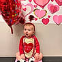 baby, toddler, valentine, heart_balloon, headband, pom_poms, beads, onesie, socks, sitting, pink, red, paper_hearts, wall_decor, indoor, cute, surprised_expression, celebration, floor, portrait