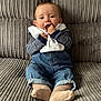 baby, child, couch, denim, overalls, sitting, striped_clothing, long_sleeves, socks, hand_in_mouth, curious, happy, face, person, indoor, soft_furniture, cozy, portrait, infant, casual