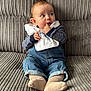 baby, child, infant, denim_overalls, striped_shirt, beige_socks, couch, ribbed_texture, curious_expression, finger_in_mouth, seated, indoors, cozy, casual_clothing, soft_lighting, portrait, cute, small_hands, young, comfortable