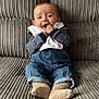 baby, child, denim_overalls, striped_shirt, couch, gray, sitting, chewing, cloth, happy, cute, infant, feet, socks, portrait, indoors, texture, casual_clothing, smiling, relaxed