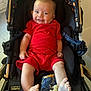 Ezra is registered to the contest to win money with this photo: baby, smiling, stroller, red_clothing, infant, child, happy, indoor, footwear, patterned_fabric, toy, legs, face, person, cute, seated, comfort, bright_colors, playful, home